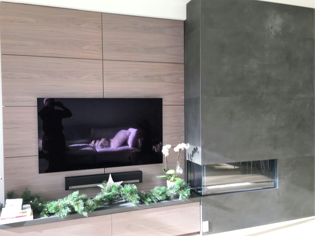 Walnut entertainment wall with recessed soundbar niche beside a dark plaster fireplace with a three-sided glass insert